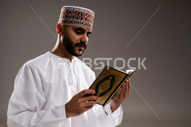 مفهوم قراءة القران، الإجتهاد في العباده والتقرب الى الله في شهر رمضان المبارك, بورتريه مقرب لرجل عربي خليجي عماني مسلم يرتدي الكمة والدشداشة يجلس على سجادة الصلاة ويقرأ المصحف الشريف, خلفية رمادية