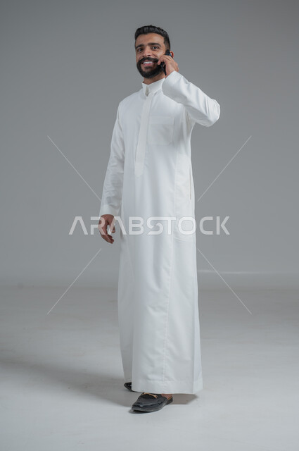 Communicating with family and friends via mobile phone, using modern and advanced devices, answering communications and calls, portrait of a Saudi Gulf Arab man wearing a traditional thobe making a phone call via mobile phone, full-length body photo, gray background