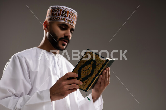 مفهوم قراءة القران، الإجتهاد في العباده والتقرب الى الله في شهر رمضان المبارك, بورتريه مقرب لرجل عربي خليجي عماني مسلم يرتدي الكمة والدشداشة يجلس على سجادة الصلاة ويقرأ المصحف الشريف, خلفية رمادية