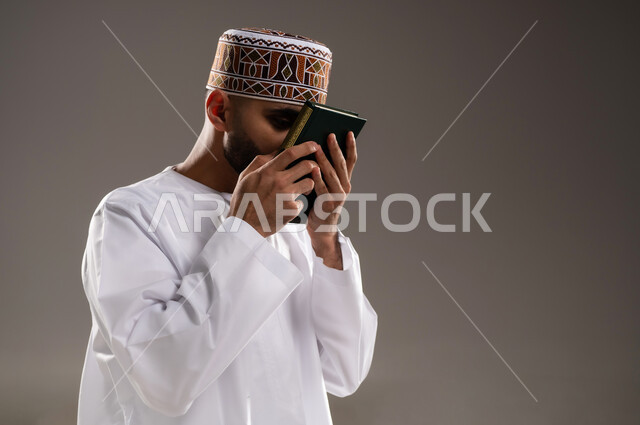 مفهوم قراءة القران، الإجتهاد في العباده والتقرب الى الله في شهر رمضان المبارك, بورتريه مقرب لرجل عربي خليجي عماني مسلم يرتدي الكمة والدشداشة يجلس على سجادة الصلاة ويقبل المصحف الشريف, خلفية رمادية