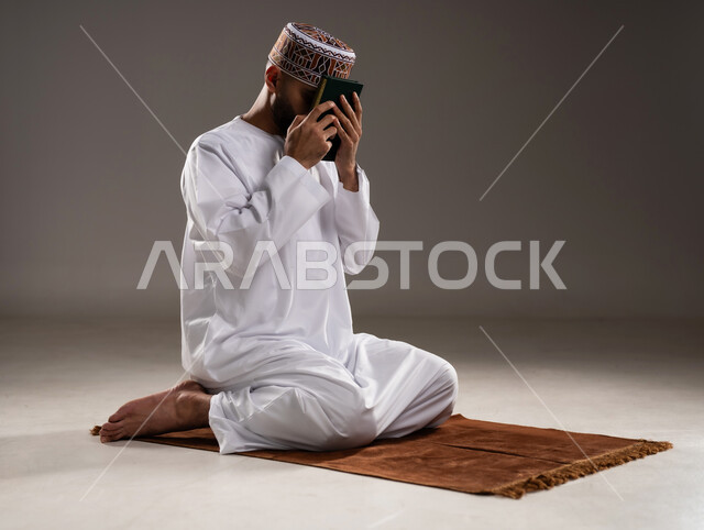 مفهوم قراءة القران، الإجتهاد في العباده والتقرب الى الله في شهر رمضان المبارك, بورتريه لرجل عربي خليجي عماني مسلم يرتدي الكمة والدشداشة يجلس على سجادة الصلاة ويقبل المصحف الشريف, خلفية رمادية