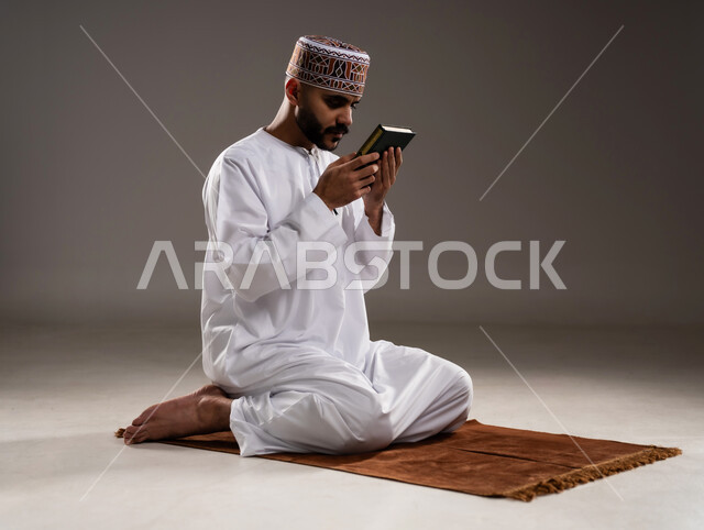مفهوم قراءة القران، الإجتهاد في العباده والتقرب الى الله في شهر رمضان المبارك, بورتريه لرجل عربي خليجي عماني مسلم يرتدي الكمة والدشداشة يجلس على سجادة الصلاة ويقبل المصحف الشريف, خلفية رمادية