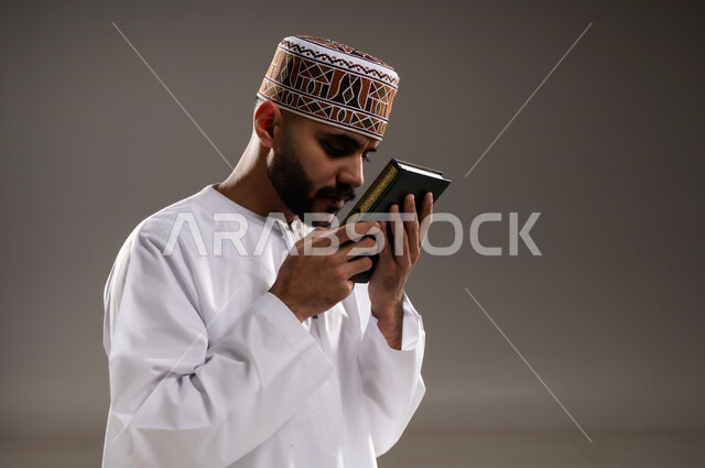مفهوم قراءة القران، الإجتهاد في العباده والتقرب الى الله في شهر رمضان المبارك, بورتريه مقرب لرجل عربي خليجي عماني مسلم يرتدي الكمة والدشداشة يجلس على سجادة الصلاة ويقبل المصحف الشريف, خلفية رمادية