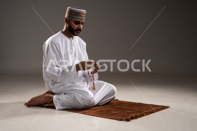 مفهوم التسبيح والتهليل، الإجتهاد في العباده والتقرب الى الله في شهر رمضان المبارك, بورتريه لرجل عربي خليجي عماني مسلم يرتدي الكمة والدشداشة يجلس على سجادة الصلاة, الإكثار من ذكر الله تعالى من خلال الإستغفار والدعاء, خلفية رمادية