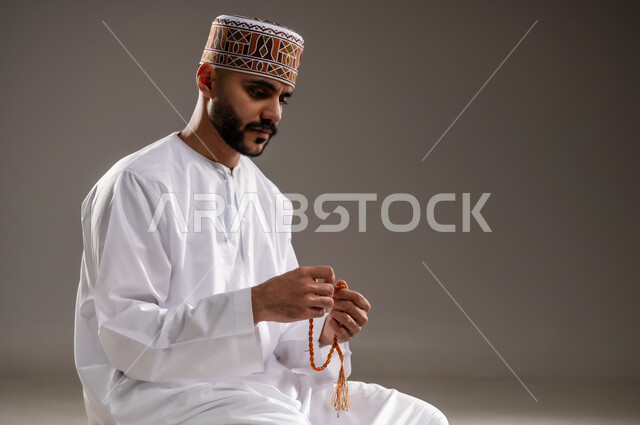 مفهوم التسبيح والتهليل، الإجتهاد في العباده والتقرب الى الله في شهر رمضان المبارك, بورتريه مقرب لرجل عربي خليجي عماني مسلم يرتدي الكمة والدشداشة يجلس على سجادة الصلاة, الإكثار من ذكر الله تعالى من خلال الإستغفار والدعاء, خلفية رمادية