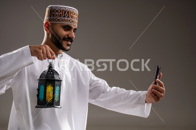مفهوم الاحتفال بقدوم المناسبات الدينية، التقاط الصور التذكارية, فوانيس اسلامية للاعياد في سلطنه عمان, الالتزام بأداء الفروض على وقتها, التقرب الى الله بالإجتهاد في العبادة والحفاظ على الصلوات، بورتريه مقرب لرجل عربي خليجي عماني مسلم يرتدي الكمة والدشداشة, الجلوس على سجادة الصلاة وحمل فانوس رمضاني وهاتف محمول، خلفية رمادية