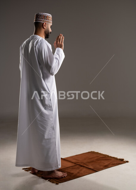 The concept of the pillars of Islam, worship and closeness to God, performing prayers on time, performing Islamic religious duties, portrait from the back of an Arab Gulf Omani Muslim man wearing a kuma and dishdasha praying, full-length body portrait, gray background
