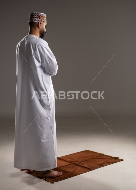 مفهوم أركان الإسلام، العبادة والتقرب إلى الله، إقامة الصلاة على وقتها، أداء الفرائض الدينية الاسلامية، بورتريه من الخلف لرجل عربي خليجي عماني مسلم يرتدي الكمة والدشداشة يصلي, صورة الجسم بالطول الكامل, خلفية رمادية