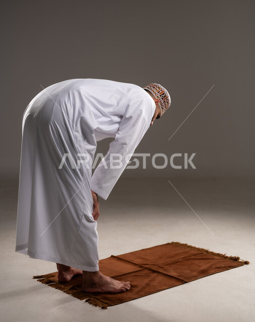 إقامة الصلوات على وقتها، مفهوم أركان الإسلام، العبادة والتقرب إلى الله، أداء الفرائض الدينية الاسلامية، بورتريه من الخلف لرجل عربي خليجي عماني مسلم يرتدي الكمة والدشداشة يركع على سجادة الصلاة, خلفية رمادية