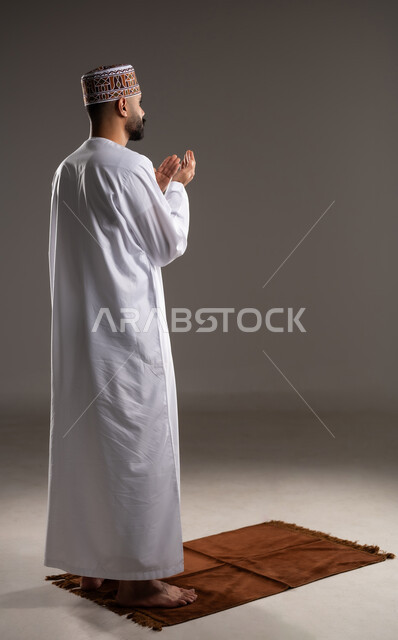 مفهوم التضرع والتعبد، الإجتهاد في العباده في شهر رمضان المبارك, بورتريه مقرب لرجل عربي خليجي عماني مسلم يرتدي الكمة والدشداشة يقف على سجادة الصلاة ويرفع يديه ويدعي, الإكثار من ذكر الله تعالى من خلال الإستغفار والدعاء, صورة الجسم بالطول الكامل, خلفية رمادية