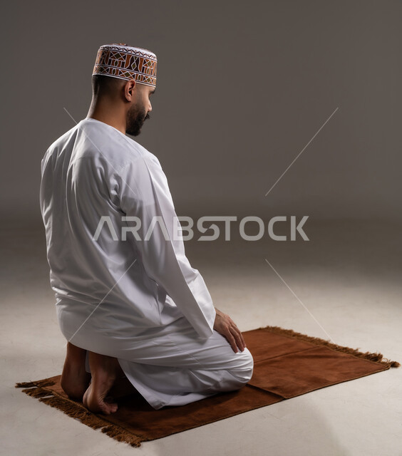 The concept of the pillars of Islam, worship and closeness to God, performing prayers on time, performing Islamic religious duties, a portrait from the back of an Arab Gulf Omani Muslim man wearing a kuma and dishdasha praying on a prayer rug, gray background