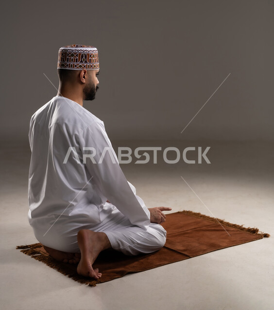 مفهوم أركان الإسلام، العبادة والتقرب إلى الله، إقامة الصلوات على وقتها، أداء الفرائض الدينية الاسلامية، بورتريه من الخلف لرجل عربي خليجي عماني مسلم يرتدي الكمة والدشداشة يصلي على سجادة الصلاة, خلفية رمادية