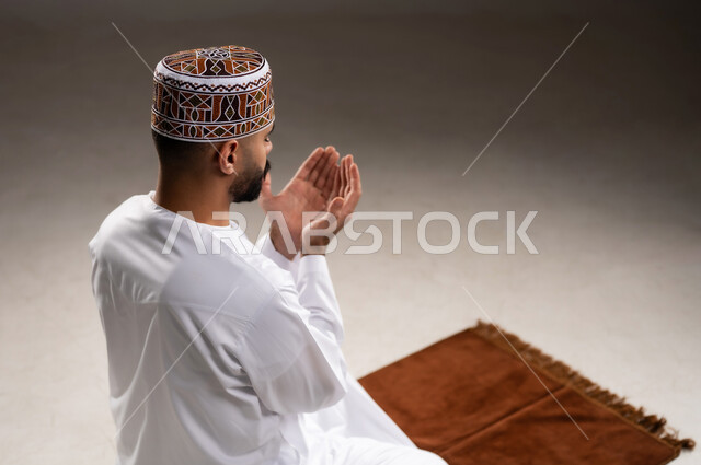 مفهوم التضرع والتعبد، الإجتهاد في العباده في شهر رمضان المبارك, بورتريه مقرب لرجل عربي خليجي عماني مسلم يرتدي الكمة والدشداشة يجلس على سجادة الصلاة ويرفع يديه ويدعي, الإكثار من ذكر الله تعالى من خلال الإستغفار والدعاء, خلفية رمادية