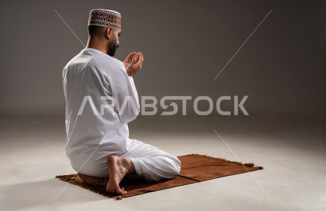 مفهوم التضرع والتعبد، الإجتهاد في العباده في شهر رمضان المبارك, بورتريه مقرب لرجل عربي خليجي عماني مسلم يرتدي الكمة والدشداشة يجلس على سجادة الصلاة ويرفع يديه ويدعي, الإكثار من ذكر الله تعالى من خلال الإستغفار والدعاء,  خلفية رمادية