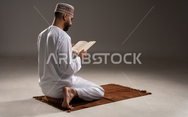 مفهوم قراءة القران، الإجتهاد في العباده والتقرب الى الله في شهر رمضان المبارك, بورتريه  لرجل عربي خليجي عماني مسلم يرتدي الكمة والدشداشة يجلس على سجادة الصلاة ويقرأ المصحف الشريف, خلفية رمادية
