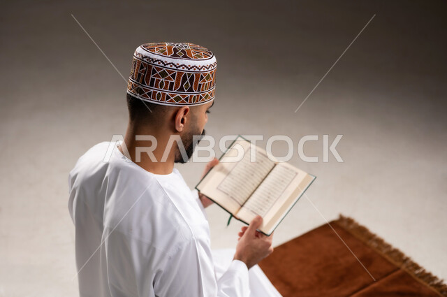 مفهوم قراءة القران، الإجتهاد في العباده والتقرب الى الله في شهر رمضان المبارك, بورتريه مقرب لرجل عربي خليجي عماني مسلم يرتدي الكمة والدشداشة يجلس على سجادة الصلاة ويقرأ المصحف الشريف, خلفية رمادية