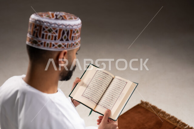 مفهوم قراءة القران، الإجتهاد في العباده والتقرب الى الله في شهر رمضان المبارك, بورتريه مقرب لرجل عربي خليجي عماني مسلم يرتدي الكمة والدشداشة يجلس على سجادة الصلاة ويقرأ المصحف الشريف, خلفية رمادية