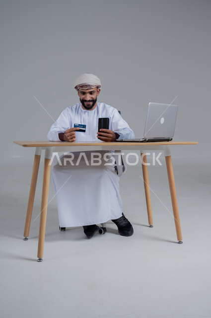 استخدام البطاقة الذكية للدفع الالكتروني، مهن ووظائف مكتبيه عمانية، بورتريه لشاب عربي خليجي عماني يرتدي الدشداشة والعمامة يجلس خلف طاولة خشبية وامامه حاسوب ويحمل بيده هاتف محمول، استخدام الأجهزة الحديثة والتقنيات المتطورة، خلفية بيضاء