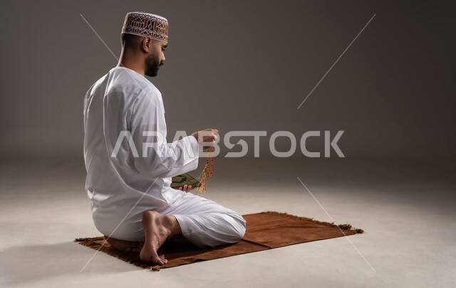 مفهوم التسبيح والتهليل وقراءة القران، الإجتهاد في العباده والتقرب الى الله في شهر رمضان المبارك, بورتريه مقرب لرجل عربي خليجي عماني مسلم يرتدي الكمة والدشداشة يجلس على سجادة الصلاة, الإكثار من ذكر الله تعالى من خلال الإستغفار والدعاء, خلفية رمادية