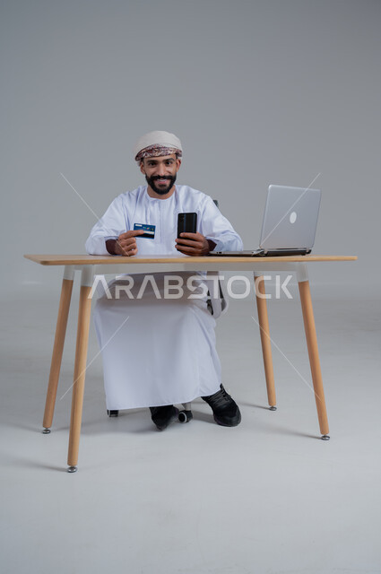 استخدام البطاقة الذكية للدفع الالكتروني، مهن ووظائف مكتبيه عمانية، بورتريه لشاب عربي خليجي عماني يرتدي الدشداشة والعمامة يجلس خلف طاولة خشبية وامامه حاسوب ويحمل بيده هاتف محمول، استخدام الأجهزة الحديثة والتقنيات المتطورة، خلفية بيضاء