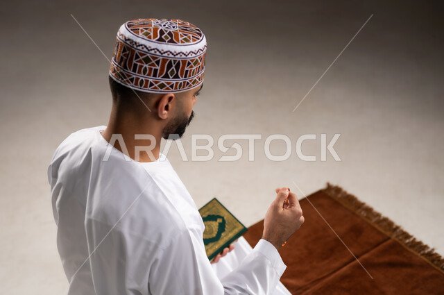 مفهوم التسبيح والتهليل وقراءة القران، الإجتهاد في العباده والتقرب الى الله في شهر رمضان المبارك, بورتريه مقرب لرجل عربي خليجي عماني مسلم يرتدي الكمة والدشداشة يجلس على سجادة الصلاة, الإكثار من ذكر الله تعالى من خلال الإستغفار والدعاء, خلفية رمادية