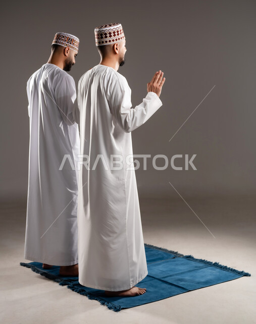مفهوم أركان الإسلام، العبادة والتقرب إلى الله، إقامة الصلاة على وقتها، أداء الفرائض الدينية الاسلامية، بورتريه من الخلف لرجلان عربيان خليجيان عمانيان مسلمان يرتديان الكمة والدشداشة يصلون صلاة الجماعة, صورة الجسم بالطول الكامل, خلفية رمادية