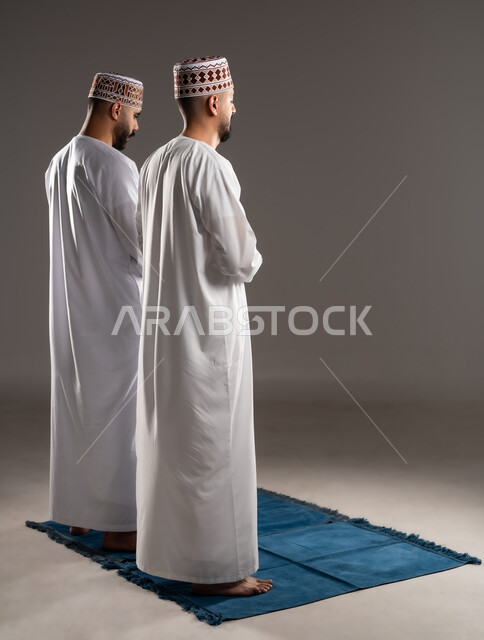 مفهوم أركان الإسلام، العبادة والتقرب إلى الله، إقامة الصلاة على وقتها، أداء الفرائض الدينية الاسلامية، بورتريه من الخلف لرجلان عربيان خليجيان عمانيان مسلمان يرتديان الكمة والدشداشة يصلون صلاة الجماعة, صورة الجسم بالطول الكامل, خلفية رمادية