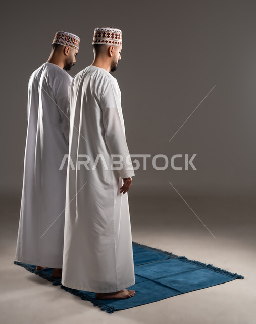 مفهوم أركان الإسلام، العبادة والتقرب إلى الله، إقامة الصلاة على وقتها، أداء الفرائض الدينية الاسلامية، بورتريه من الخلف لرجلان عربيان خليجيان عمانيان مسلمان يرتديان الكمة والدشداشة يصلون صلاة الجماعة, صورة الجسم بالطول الكامل, خلفية رمادية