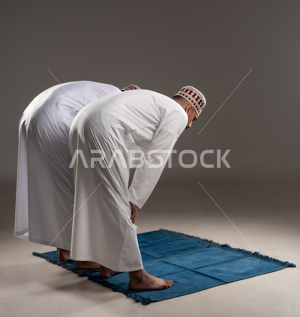 إقامة الصلوات على وقتها، العبادة والتقرب إلى الله، أداء الفرائض الدينية الاسلامية، مفهوم أركان الإسلام، بورتريه من الخلف لرجلان عربيان خليجيان عمانيان مسلمان يرتديان الكمة والدشداشة ويركعان على سجادة الصلاة, خلفية رمادية