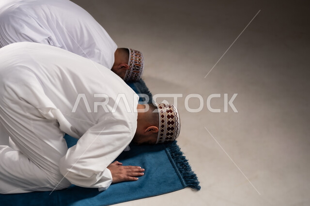 إقامة الصلوات على وقتها، العبادة والتقرب إلى الله، أداء الفرائض الدينية الاسلامية، مفهوم أركان الإسلام، بورتريه من الجانب لرجلان عربيان خليجيان عمانيان مسلمان يرتديان الكمة والدشداشة ويسجدان على سجادة الصلاة, خلفية رمادية