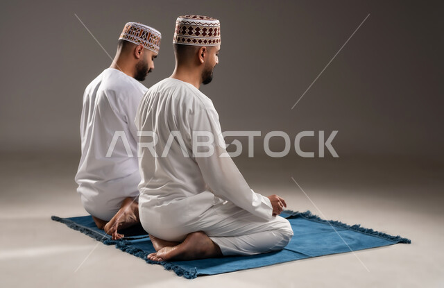 العبادة والتقرب إلى الله، إقامة الصلوات على وقتها، أداء الفرائض الدينية الاسلامية، مفهوم أركان الإسلام، بورتريه من الخلف  لرجلان عربيان خليجيان عمانيان مسلمان يرتديان الكمة والدشداشة ويجلسان على سجادة الصلاة, خلفية رمادية