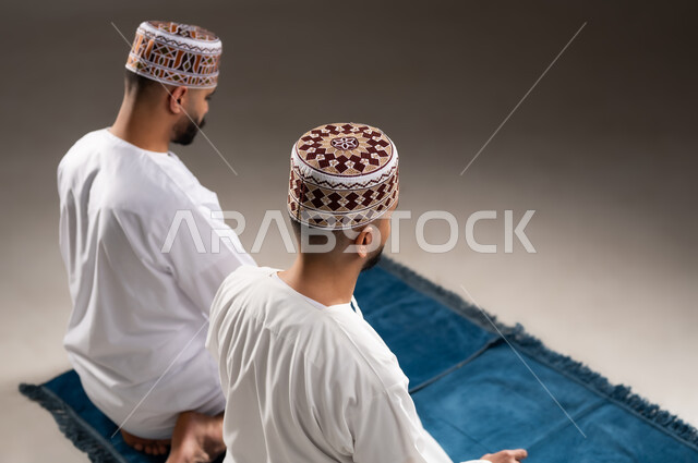 العبادة والتقرب إلى الله، إقامة الصلوات على وقتها، أداء الفرائض الدينية الاسلامية، مفهوم أركان الإسلام، بورتريه مقرب من الخلف  لرجلان عربيان خليجيان عمانيان مسلمان يرتديان الكمة والدشداشة ويجلسان على سجادة الصلاة, خلفية رمادية