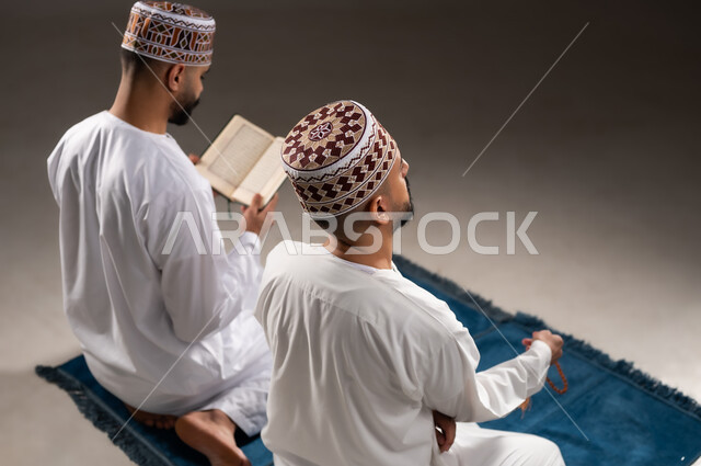 مفهوم التسبيح والتهليل وقراءة القران، الإجتهاد في العباده والتقرب الى الله في شهر رمضان المبارك, بورتريه مقرب من الخلف لرجلان عربيان خليجيان عمانيان مسلمان يرتديان الكمة والدشداشة يجلسان على سجادة الصلاة, الإكثار من ذكر الله تعالى من خلال الإستغفار والدعاء, خلفية رمادية