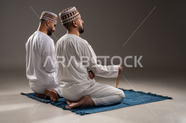 مفهوم التسبيح والتهليل وقراءة القران، الإجتهاد في العباده والتقرب الى الله في شهر رمضان المبارك, بورتريه من الخلف لرجلان عربيان خليجيان عمانيان مسلمان يرتديان الكمة والدشداشة يجلسان على سجادة الصلاة, الإكثار من ذكر الله تعالى من خلال الإستغفار والدعاء, خلفية رمادية