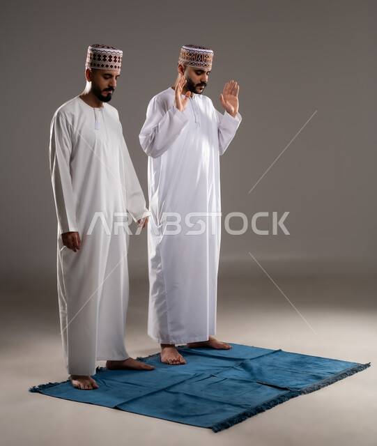 العبادة والتقرب إلى الله، إقامة الصلاة على وقتها، أداء الفرائض الدينية الاسلامية، مفهوم أركان الإسلام، بورتريه لرجلان عربيان خليجيان عمانيان مسلمان يرتديان الكمة والدشداشة  يصلون صلاة الجماعة, صورة الجسم بالطول الكامل, خلفية رمادية