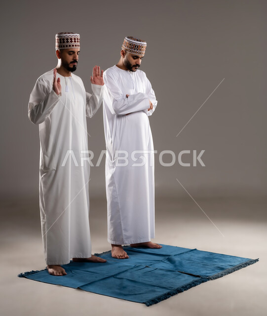 العبادة والتقرب إلى الله، إقامة الصلاة على وقتها، أداء الفرائض الدينية الاسلامية، مفهوم أركان الإسلام، بورتريه لرجلان عربيان خليجيان عمانيان مسلمان يرتديان الكمة والدشداشة  يصلون صلاة الجماعة, صورة الجسم بالطول الكامل, خلفية رمادية