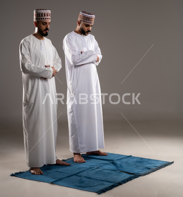 العبادة والتقرب إلى الله، إقامة الصلاة على وقتها، أداء الفرائض الدينية الاسلامية، مفهوم أركان الإسلام، بورتريه لرجلان عربيان خليجيان عمانيان مسلمان يرتديان الكمة والدشداشة  يصلون صلاة الجماعة, صورة الجسم بالطول الكامل, خلفية رمادية