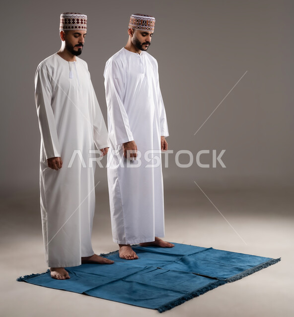 العبادة والتقرب إلى الله، إقامة الصلاة على وقتها، أداء الفرائض الدينية الاسلامية، مفهوم أركان الإسلام، بورتريه لرجلان عربيان خليجيان عمانيان مسلمان يرتديان الكمة والدشداشة  يصلون صلاة الجماعة, صورة الجسم بالطول الكامل, خلفية رمادية
