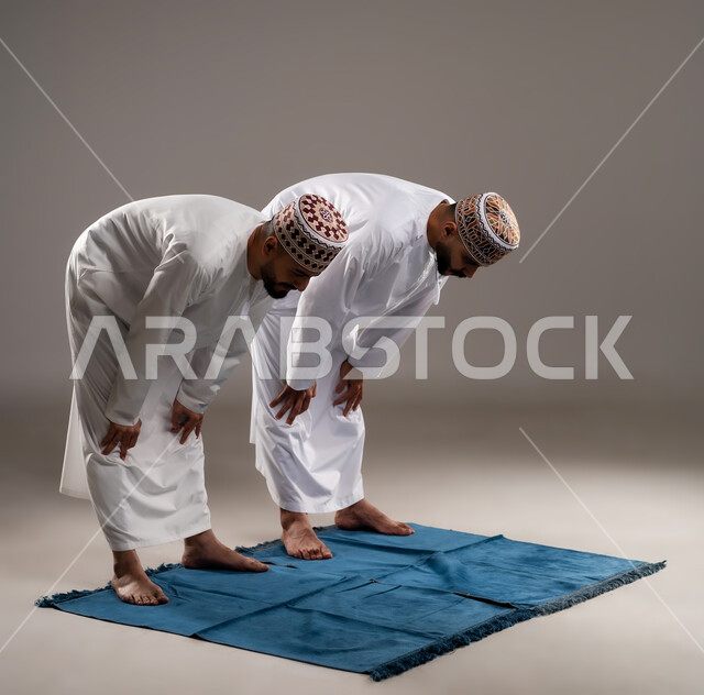 إقامة الصلوات على وقتها، العبادة والتقرب إلى الله، أداء الفرائض الدينية الاسلامية، مفهوم أركان الإسلام، بورتريه لرجلان عربيان خليجيان عمانيان مسلمان يرتديان الكمة والدشداشة ويركعان على سجادة الصلاة, خلفية رمادية