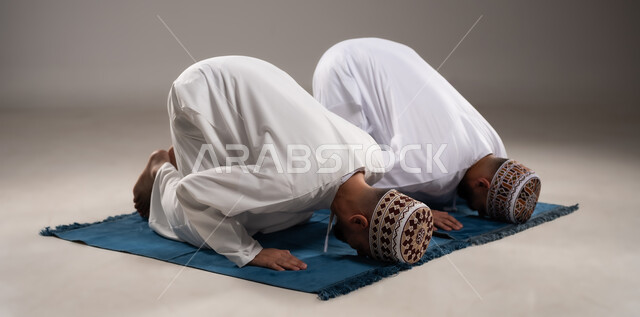 العبادة والتقرب إلى الله، إقامة الصلوات على وقتها، أداء الفرائض الدينية الاسلامية، مفهوم أركان الإسلام، بورتريه لرجلان عربيان خليجيان عمانيان مسلمان يرتديان الكمة والدشداشة ويسجدان على سجادة الصلاة, خلفية رمادية