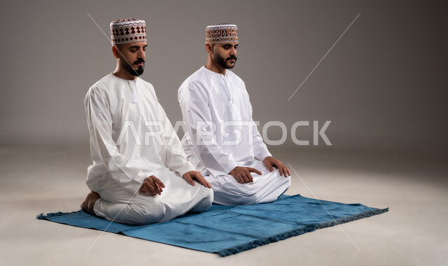 العبادة والتقرب إلى الله، إقامة الصلوات على وقتها، أداء الفرائض الدينية الاسلامية، مفهوم أركان الإسلام، بورتريه لرجلان عربيان خليجيان عمانيان مسلمان يرتديان الكمة والدشداشة ويجلسان على سجادة الصلاة, خلفية رمادية