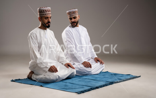 العبادة والتقرب إلى الله، إقامة الصلوات على وقتها، أداء الفرائض الدينية الاسلامية، مفهوم أركان الإسلام، بورتريه لرجلان عربيان خليجيان عمانيان مسلمان يرتديان الكمة والدشداشة ويجلسان على سجادة الصلاة, خلفية رمادية