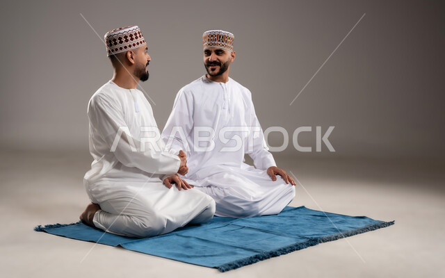 مفهوم القاء التحية، بورتريه لرجلان عربيان خليجيان عمانيان مسلمان يرتديان الكمة والدشداشة يجلسان على سجادة الصلاة ويتبادلون السلام, أداء فريضة الصلاه داخل المسجد، خلفية رمادية