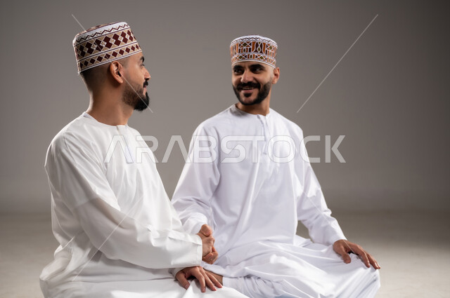 مفهوم القاء التحية، بورتريه لرجلان عربيان خليجيان عمانيان مسلمان يرتديان الكمة والدشداشة يجلسان على سجادة الصلاة ويتبادلون السلام, أداء فريضة الصلاه داخل المسجد، خلفية رمادية
