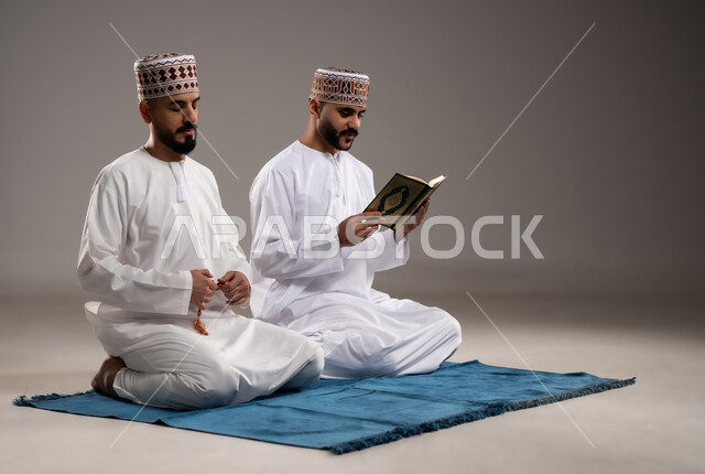مفهوم التسبيح والتهليل وقراءة القران، الإجتهاد في العباده والتقرب الى الله في شهر رمضان المبارك, بورتريه لرجلان عربيان خليجيان عمانيان مسلمان يرتديان الكمة والدشداشة يجلسان على سجادة الصلاة, الإكثار من ذكر الله تعالى من خلال الإستغفار والدعاء, خلفية رمادية