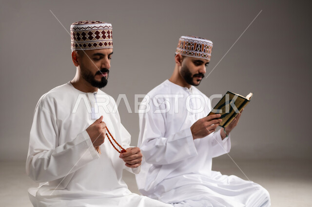 مفهوم التسبيح والتهليل وقراءة القران، الإجتهاد في العباده والتقرب الى الله في شهر رمضان المبارك, بورتريه مقرب لرجلان عربيان خليجيان عمانيان مسلمان يرتديان الكمة والدشداشة يجلسان على سجادة الصلاة, الإكثار من ذكر الله تعالى من خلال الإستغفار والدعاء, خلفية رمادية