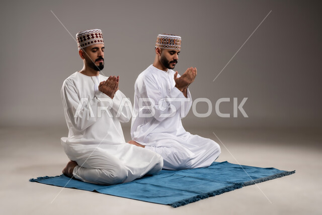 مفهوم التضرع الى الله، الإجتهاد في العباده في شهر رمضان المبارك, بورتريه لرجلان عربيان خليجيان عمانيان مسلمان يرتديان الكمة والدشداشة يجلسان على سجادة الصلاة ويرفعان ايديهم ويدعيان, الإكثار من ذكر الله تعالى من خلال الإستغفار والدعاء, خلفية رمادية