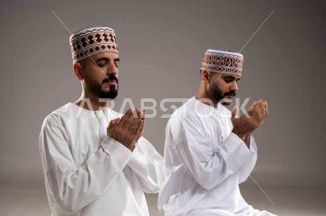 مفهوم التضرع الى الله، الإجتهاد في العباده في شهر رمضان المبارك, بورتريه مقرب لرجلان عربيان خليجيان عمانيان مسلمان يرتديان الكمة والدشداشة يجلسان على سجادة الصلاة ويرفعان ايديهم ويدعيان, الإكثار من ذكر الله تعالى من خلال الإستغفار والدعاء, خلفية رمادية