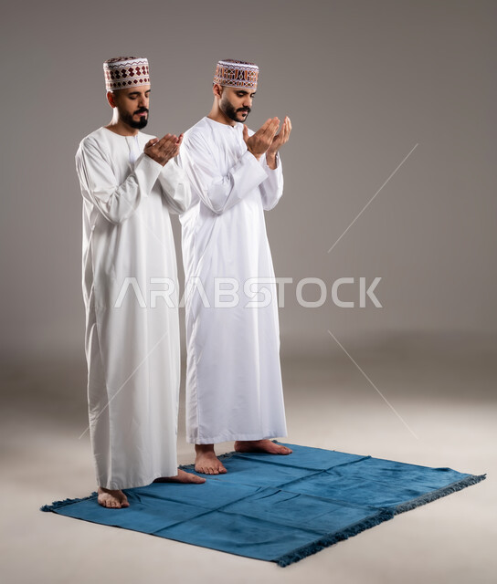 مفهوم التضرع والتعبد، الإجتهاد في العباده في شهر رمضان المبارك, بورتريه مقرب لرجلان عربيان خليجيان عمانيان مسلمان يرتديان الكمة والدشداشة يقفان على سجادة الصلاة ويرفعان ايديهم ويدعيان, الإكثار من ذكر الله تعالى من خلال الإستغفار والدعاء, خلفية رمادية