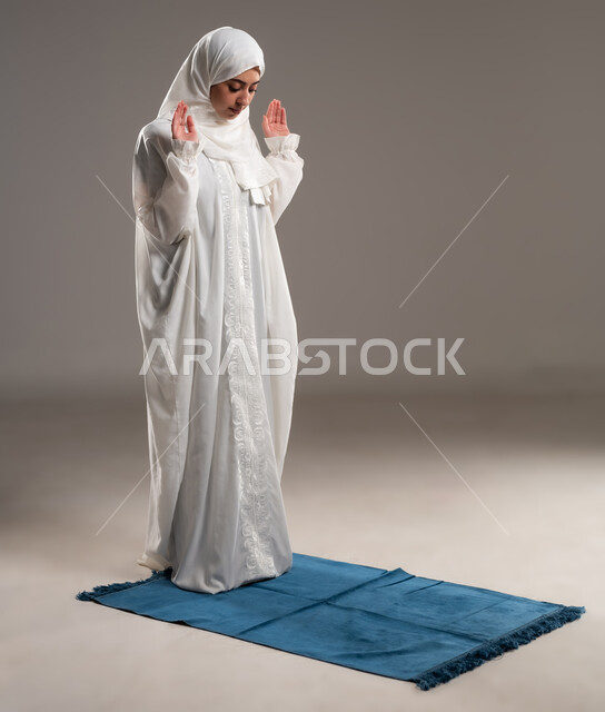 ايماءات تدل على الخشوع في الصلاة, أداء الفروض على وقتها, التقرب الى الله من خلال الإجتهاد في العبادة والحفاظ على الصلوات، بورتريه  لإمرأة عربية عمانية خليجية ترتدي الحجاب والعباءة البيضاء تقف على سجادة الصلاة, صورة الجسم بالطول الكامل, خلفية رمادية